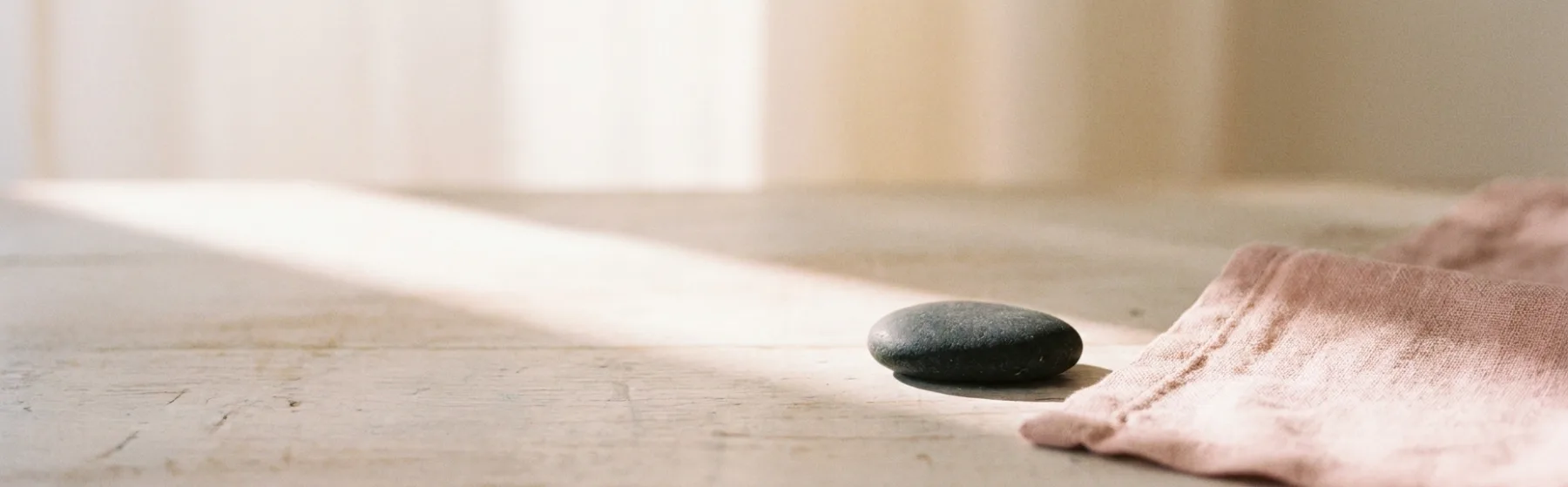 A pale wood surface with warm rose light falling across it, a smooth river stone and a soft blush linen edge